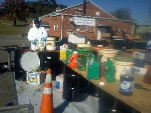 Five gallon buckets of chemicals collected on Hazardous Waste Day sitting on a table. A worker with a white haz-mat suite is disposing of some of the chemicals.